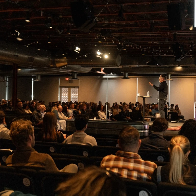 A view from the back of a crowded auditorium as a pastor speaks on stage during a church service. The audience is seated and looking towards the stage.